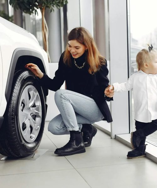 Full length of cheerful smiling mother in jeans holding hands with cute little girl and looking at car wheel in car showroom in daylight